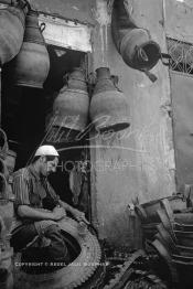 Image du Maroc Professionnelle de  Un Homme découpe un pneu de camion pour le transformer  en différents objets dont quelques uns sont accrochés au dessus. Plusieurs ateliers existaient dans rue situés derrière le cinéma Mabrouka où quelques artisans transformaient des pneus usagés en sceau pour hammam, cruche ou jarre que les paysans utilisaient pour faire sortir l’eau des puits. A cette époque le recyclage des pneus faisait partie intégrante du paysage social au Maroc, presque tous les hammams ont utilisés ces sceaux en caoutchouc à la place de ceux en bois, à cause de leur prix exorbitant et surtout qui se brisaient souvent par les lancements des ouvriers aux heures du nettoyage, Mercredi 18 Mai 1988. (Photo / Abdeljalil Bounhar)